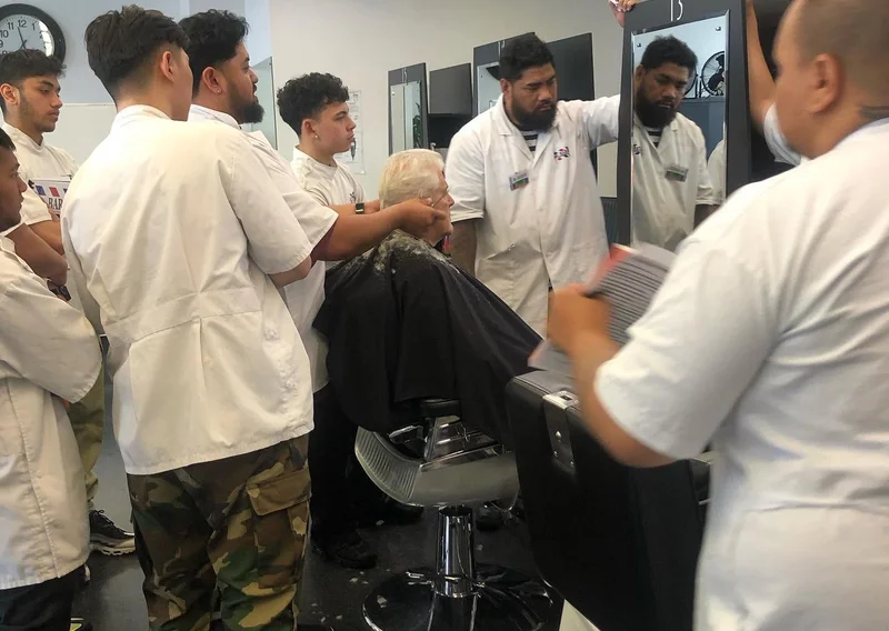 Group of trainee barbers in white coats gathered around an elderly client getting a haircut